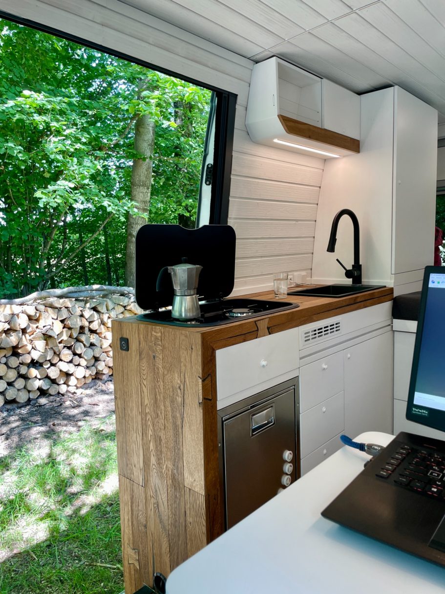 Küche in einem modernen und gemütlichen Camper Van mit Holzregal und Blick ins Grüne.